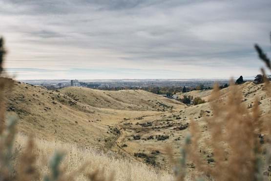 View of downtown Boise from the foothills. Features grassy hills with dirt trail and blue sky view.